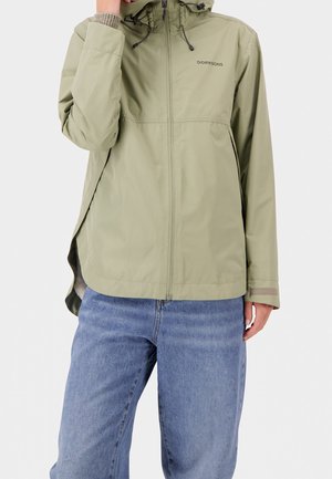 Persoon draagt een lichtgroene Didriksons-jas met capuchon en ritssluiting aan de voorkant, en blauwe jeans, met de hand dichtbij het gezicht en de andere langs het lichaam.