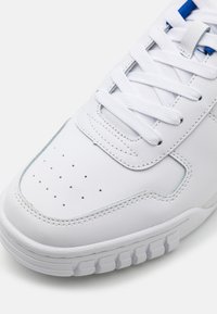 Baltos sportinės bateliai su lygiu sintetiniu paviršiumi, perforuota nosimi, plokščiais baltais raišteliais ir tekstūruotu guminio pado paviršiumi, turintys šviesiai mėlynus akcentus.