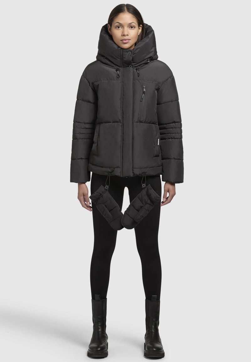 Giacca a vento nera con grande cappuccio, tasca sul petto con zip e coulisse elastiche all'orlo; abbinata a scaldamani coordinati.