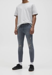 Camisa corta de manga gris combinada con jeans oscuros gris claro de corte ajustado y ligeramente desgastados, y zapatillas negras con suela texturizada.