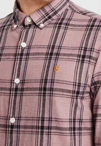 Bawełniana koszula w kratę w kolorach fioletowym i czarnym, z kołnierzykiem na guziki, białymi guzikami oraz małym haftowanym logo na piersi.