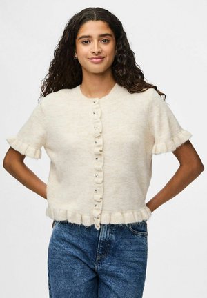 Kortärmad krämfärgad cardigan med texturerad yta, volangkanter och silverknappar framtill. Matchad med blå denim.