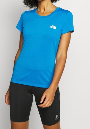 Femme portant un t-shirt North Face bleu vif à manches courtes et un short de cyclisme noir Odlo jusqu'à mi-cuisse sur un fond uni.