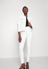 Jaqueta de ganga branca e calças brancas justas com fechos de correr ao nível do tornozelo, combinadas com uma blusa preta e sandálias pretas com tiras. Textura suave.