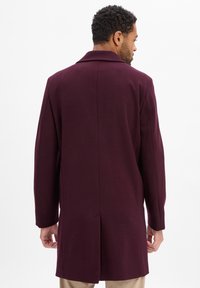 Bordeauxrode wollen overjas met een klassieke kentkraag, lange mouwen en een achterventiel voor bewegingsvrijheid. Strak, minimalistisch ontwerp.