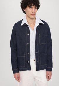 Mørkeblå denimjakke med kontrasterende hvite sømmer, har to frontlommer og trykknapper. Brukt over en stripete skjorte.