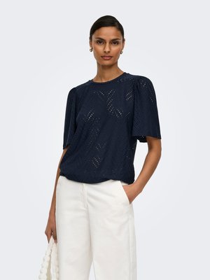 Vrouw met een marineblauw gestructureerd topje met korte mouwen, witte broek, gouden hoepeloorbellen, die een witte handtas met kralen vasthoudt, staand tegen een effen achtergrond.