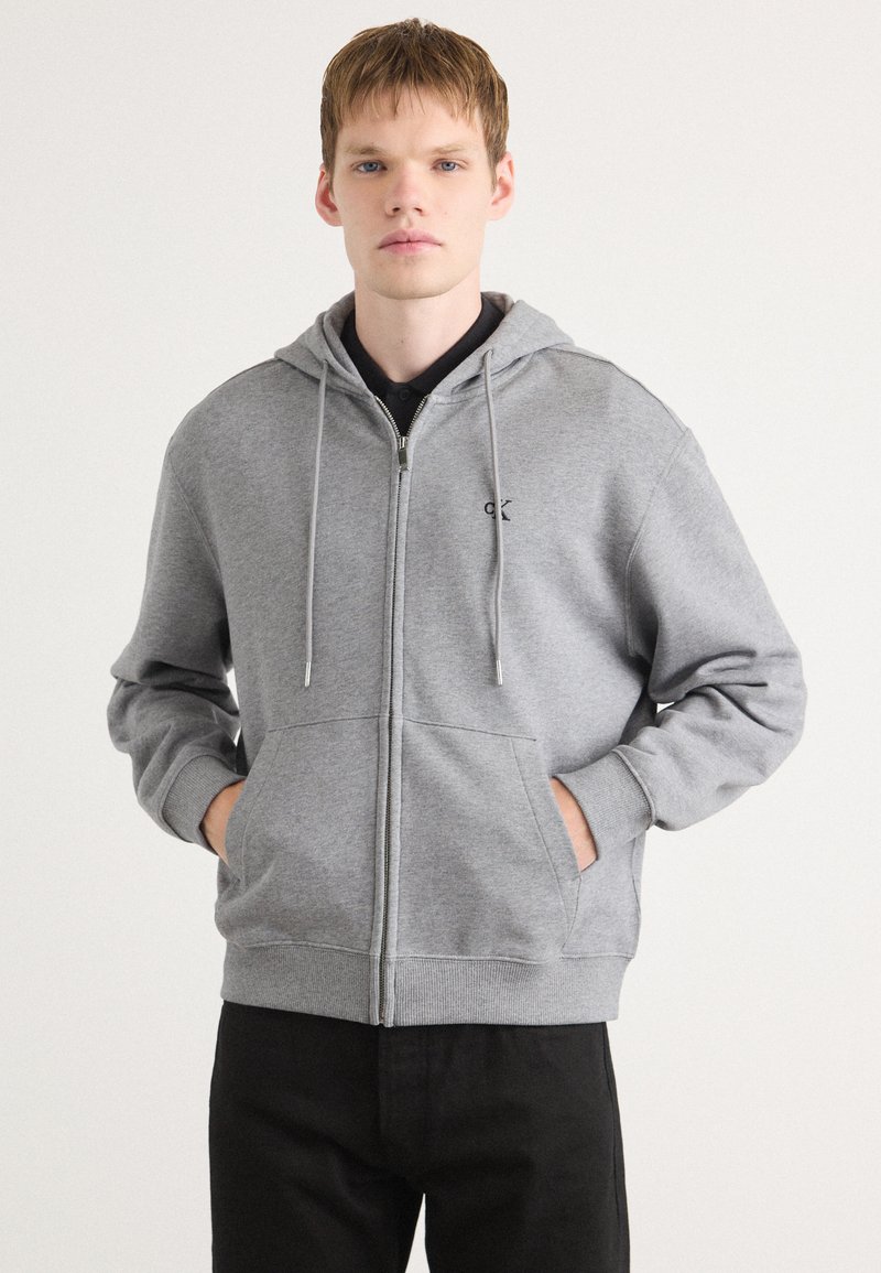 Grijze zip-up hoodie van zachte stof, met een voorzak, verstelbare trekkoordkap en een klein logo op de linkerkant van de borst.