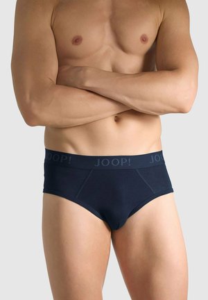 Hombre musculoso con los brazos cruzados, vistiendo calzoncillos marca Joop! azul marino, sobre un fondo liso de color gris claro.