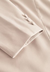 Beige kohandatud jakivars, millel on sile kangast tekstuur ja kolm ümmargust kangast kattega nuppu manseti ääres.
