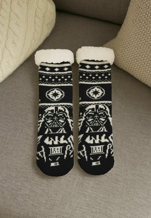 Schwarz-weiß gemusterte Wintersocken mit Darth Vader Design und Fleece-Futter, auf einer grauen Stoffoberfläche zwischen beigen strukturierten Kissen platziert.