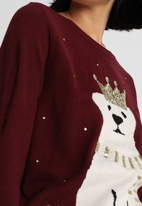 Maglione borgogna con design di un orso polare bianco, corona dorata e dettagli in paillettes. Texture morbida, scollo rotondo e dettagli giocosi.
