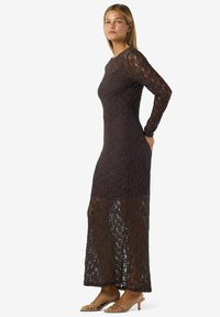Dlouhé, přiléhavé šaty z tmavě hnědé krajky, s dlouhými rukávy a poloprůhledným spodním dílem. Doplněné jednoduchým výstřihem a štíhlou siluetou.