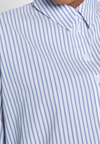 Blå och vit randig skjorta med knäppkrage, med vertikala ränder och en slät tygtextur. Fem knappar synliga på knäppningen.