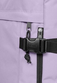 Lila tygryggsäck med dubbla dragkedjor, en svart plasthake och en snörning som accent. Stadig textur med förstärkt sömnad.