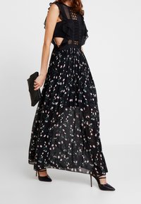 Maxi šaty s černým volánkovým topem s jemnými výřezy a splývavou plisovanou sukní zdobenou růžovými květinovými motivy na černém pozadí.