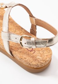 Metallinen kultainen sandaali, jossa on korkkijalkapeite. Varusteena metallinen hihna ja kultainen solkiyksityiskohta. Tasapohjainen muotoilu.