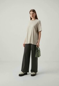 Jovem mulher a usar uma t-shirt oversized bege, calças largas de riscas verticais escuras, sapatos de plataforma brancos e um colar colorido, segurando uma pequena bolsa verde.