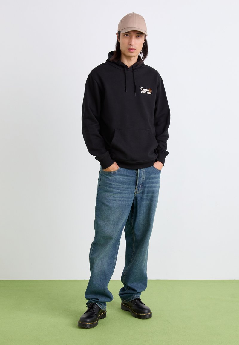 Felpa nera con tasca frontale e logo, jeans larghi blu chiaro e scarpe platform nere. Il modello indossa un cappellino beige. Sfondo verde a terra.