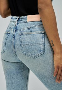 Jeans de mezclilla azul claro con un diseño ajustado, que presentan un parche de logo de cuero en la parte trasera, dos bolsillos traseros y detalles de costura visibles.