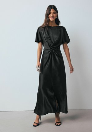 Black satin maxi dress with short flutter sleeves, knotted front detail, and side slits. Model wears black strappy heels and has silver-painted hand.