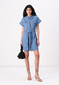 Denim srajčna obleka s kratkimi rokavi, gumbnim zapiranjem, pasom in dvema žepoma na prsih. V kombinaciji s črno torbico in sandali z vezalkami.