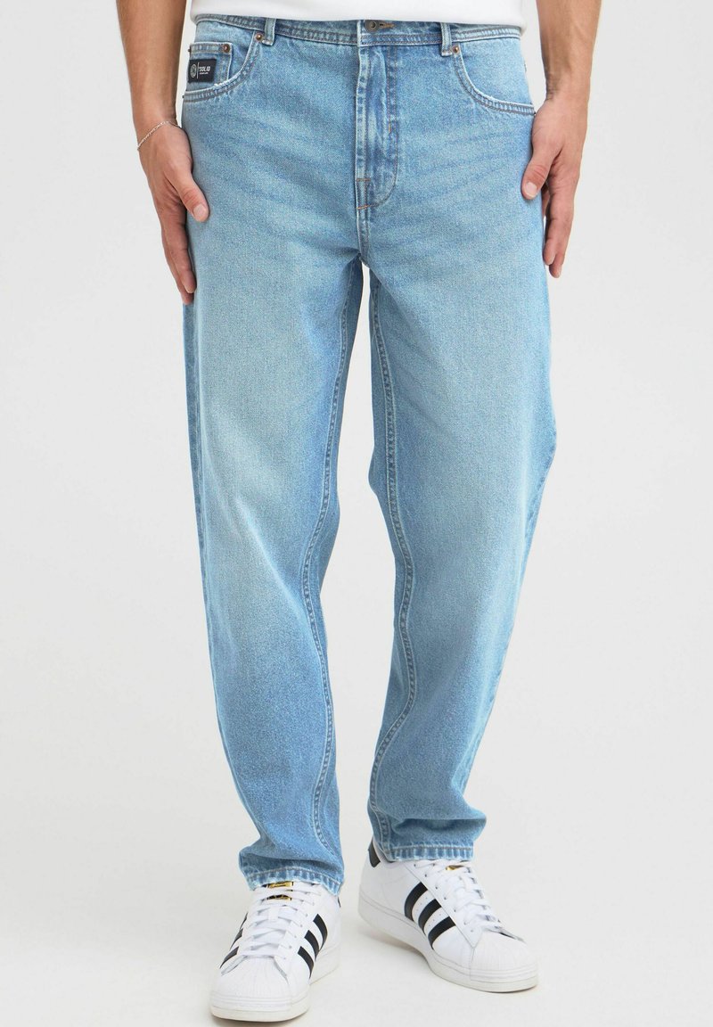 Jeans de denim azul claro con un corte relajado, que cuentan con cinco bolsillos, una cremallera de botón y un ligero desgaste, combinados con zapatillas blancas.