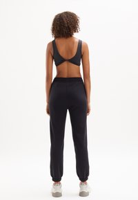 Zwarte sportbeha met open rugontwerp, gecombineerd met zwarte joggingsbroek met elastische boorden en tailleband. Model draagt witte sneakers.