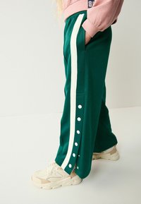 Pantalons de sport verts avec des rayures blanches sur le côté et des boutons-pression le long de la jambe, fabriqués en tissu lisse. Portés avec des baskets beiges.