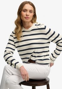 Kremowy sweter z granatowymi poziomymi paskami, okrągłym dekoltem i guzikami na ramionach. Noszony z białymi spodniami, siedzący na ciemnym drewnianym stołku.