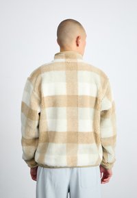 Fleece jakna v bež in beli karirasti vzorec, z visokim ovratnikom in sproščenim krojem. Mehka, teksturirana tkanina z spušenimi rameni.