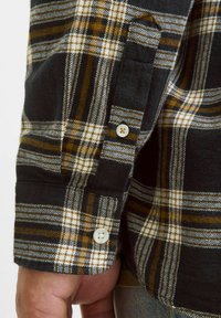 Sort og brun skotskternet flannel-skjorte med hvide knapper ved manchetterne, som har en blød tekstur og kontrasterende ternede mønstre.