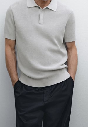 Homme portant un polo à manches courtes texturé gris clair avec col, associé à un pantalon noir ample, les mains dans les poches.