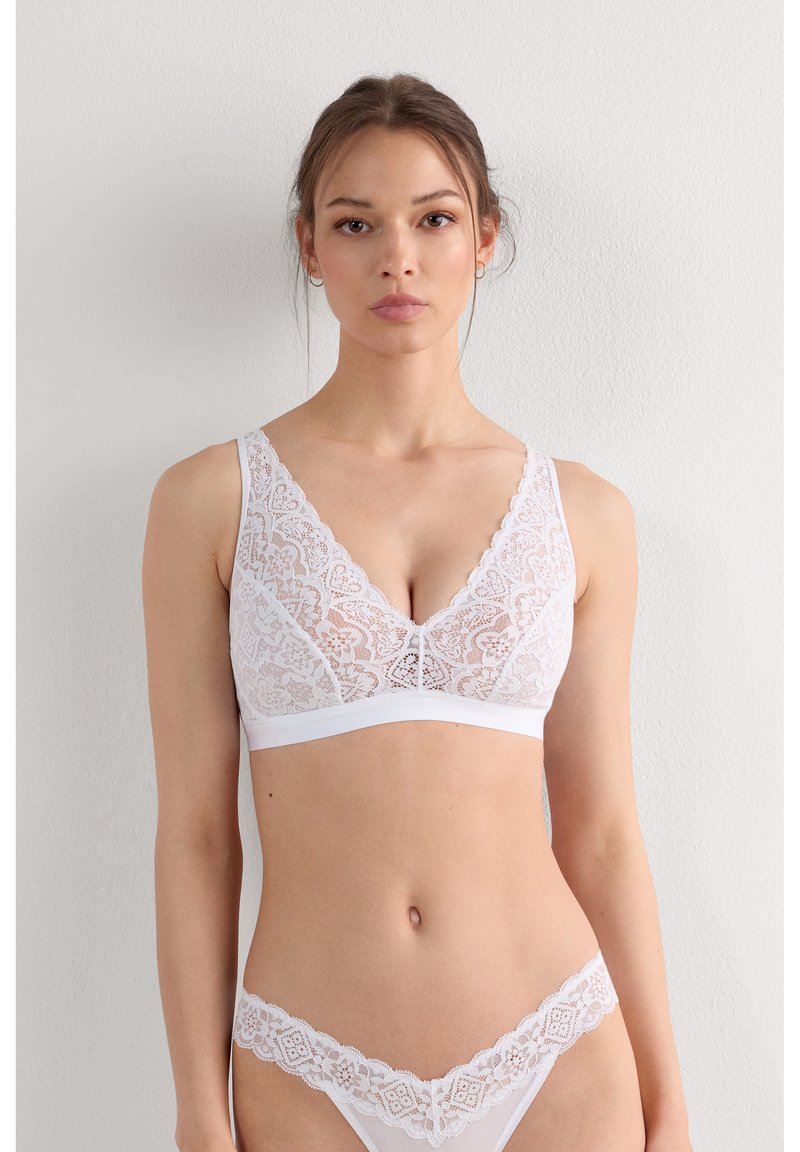 Jonge vrouw draagt bijpassende witte kanten bh en slip, staat tegen een effen lichtgekleurde muur en kijkt vooruit met een neutrale uitdrukking.