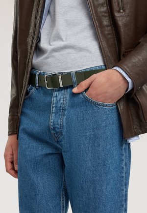 Man wearing blue jeans, gray shirt, brown leather jacket, and green belt with silver buckle, hand in jeans pocket.