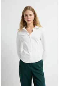 Camicia bianca a maniche lunghe aderente con colletto classico e chiusura a bottoni, caratterizzata da un tessuto texturizzato e una finitura liscia. Abbinata a pantaloni verde scuro.