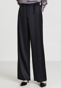 Person, die hochgeschnittene, weite schwarze Satin-Hosen mit seitlichen Taschen und schwarzen spitzen Kitten-Heels trägt, steht auf einem hellen Boden.