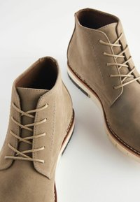 Stivaletti alla caviglia in suede marrone chiaro con punta arrotondata, suola bianca sporca e lacci beige. Presenta dettagli di cucitura lungo le cuciture.