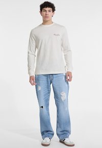 Long-sleeve cream t-shirt with logo, paired with light blue distressed wide-leg jeans and white sneakers.