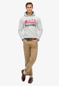 Szara bluza z kapturem z logo "Superdry" i grafiką palmy, w zestawieniu z beżowymi chinosami i burgundowymi butami. Teksturowany materiał, casualowy design.