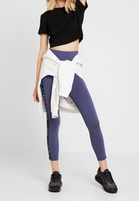 Czarny krótki t-shirt, fioletowe legginsy z bocznym logo, biały sweter związany w talii i czarne buty sportowe, o gładkiej fakturze.