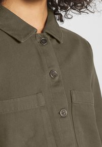 Olivově zelená košile s bundovým designem vyrobená z hladké tkaniny. Má límeček, přední zapínání na knoflíky a dvě náprsní kapsy.