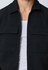 Veste noire zippée en tissu texturé, avec un col, deux poches avant et un design léger. Portée sur une chemise blanche.