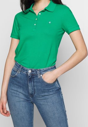 Nainen, joka pukee kirkkaan vihreän, lyhythihaisen polopaidan korkeavyötäröisten sinisten farkkujen sisään, toinen käsi taskussa.