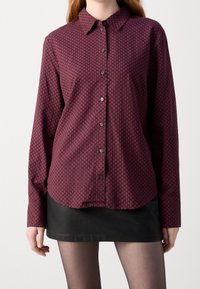 Camisa abotonada burdeos con un patrón geométrico, de mangas largas y con un collar clásico. Lleva puesta sobre una falda negra. Textura de tela suave.