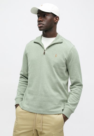 Ανοιχτό πράσινο φούτερ με φερμουάρ και μικρό κεντημένο λογότυπο, συνδυασμένο με λευκή μπλούζα και μπεζ παντελόνι; μαλακό ύφασμα και άνετη εφαρμογή.