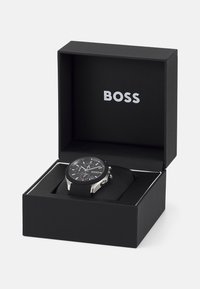 Black wristwatch in a black box with the brand name "BOSS." Features a silver case, black dial, and rubber strap with visible chronograph details.