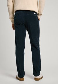 Navy corduroy pants with a straight-leg cut, featuring two back pockets and a waistband label, paired with light-colored shoes.