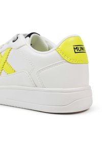 Zapatilla blanca con parte superior de cuero texturizado, que presenta un acento amarillo y un logo en el talón, ojales negros y suela de goma.