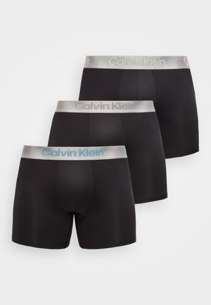Tre boxer Calvin Klein neri con elastici in vita argentati, due con logo grigio e uno con logo blu, mostrati su sfondo bianco.